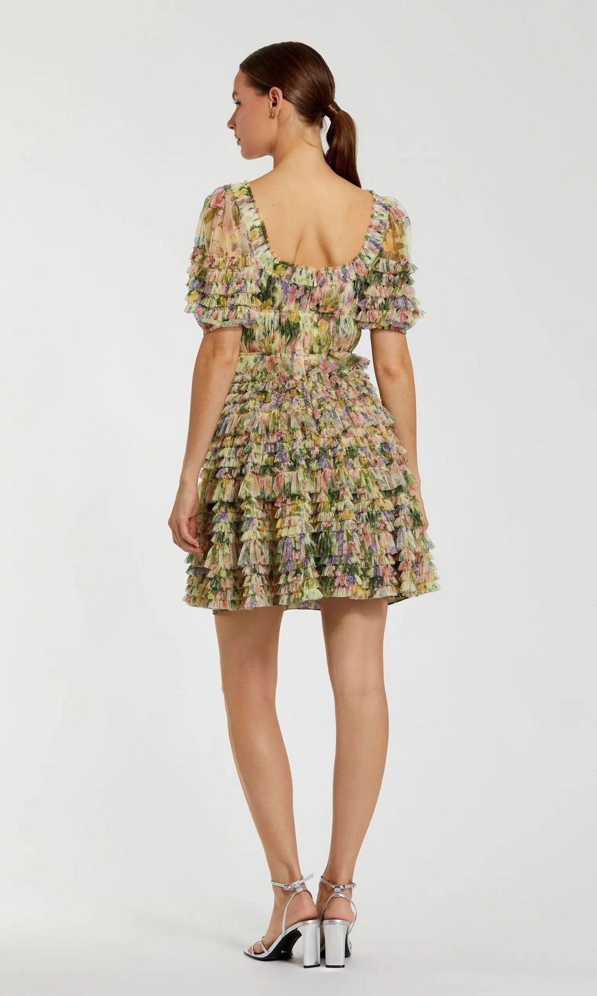 A woman is shown from behind, wearing the Mac Duggal 8327 Short Party Dress featuring a floral print with ruffled layers and puffed sleeves. She pairs it with silver sandals, and her hair is tied back in a low ponytail. The background is plain and light-colored.